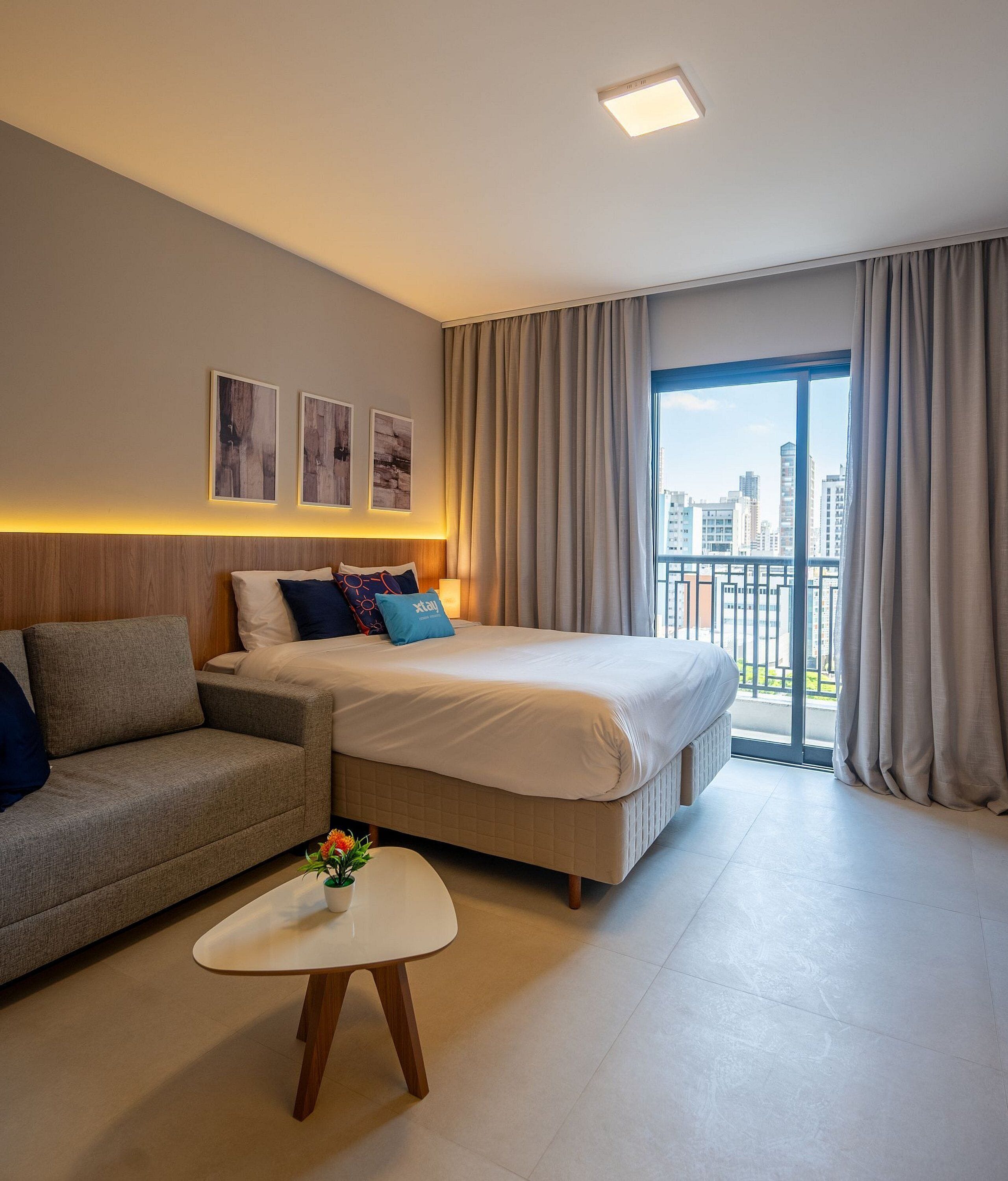 A modern hotel room with a bed, a sofa, a small table with a flower, and a city view through large windows.