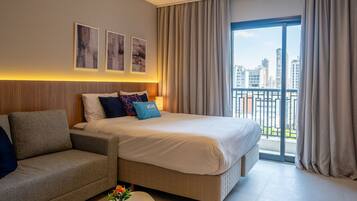 A modern hotel room with a bed, a sofa, a small table with a flower, and a city view through large windows.
