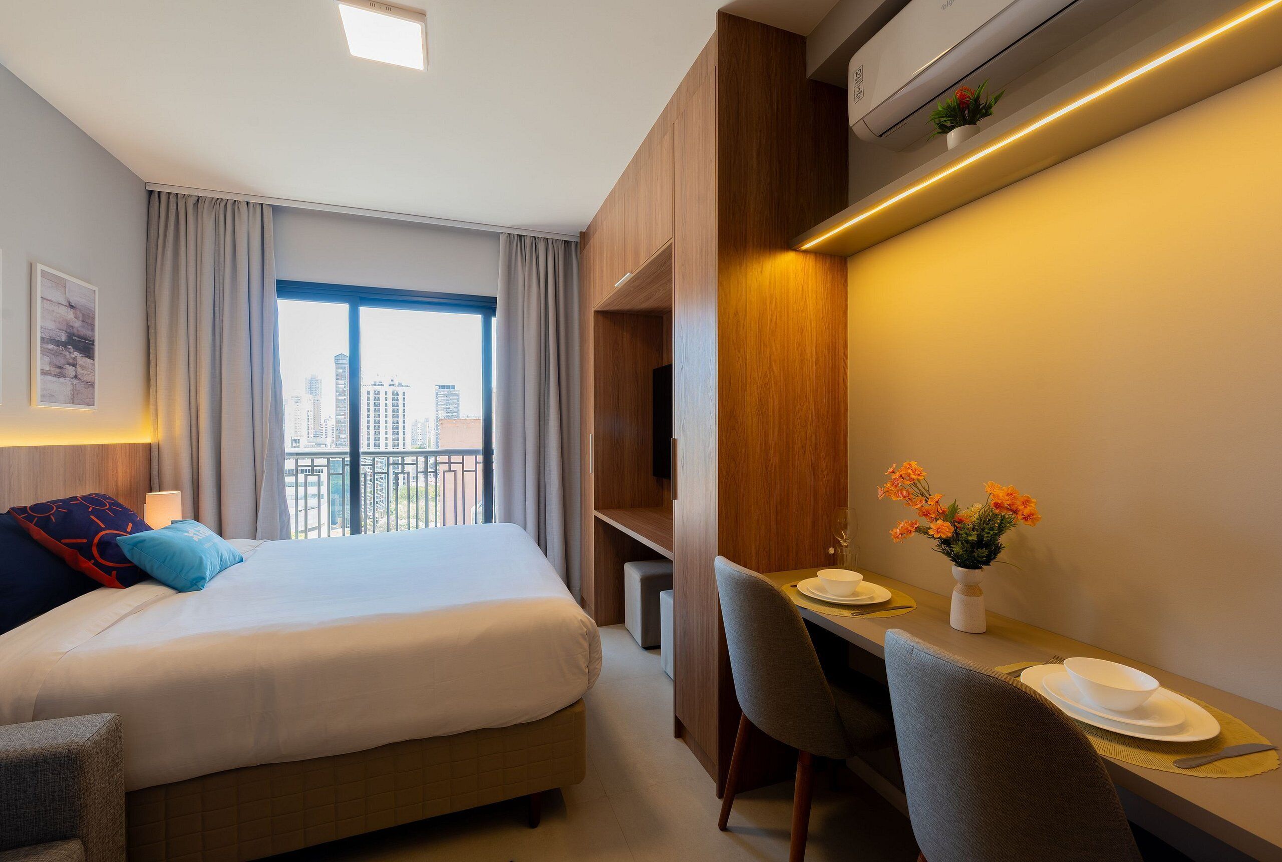 A hotel room with a bed, a desk with a chair, a small table with a vase of flowers, and a view of the city through large windows.