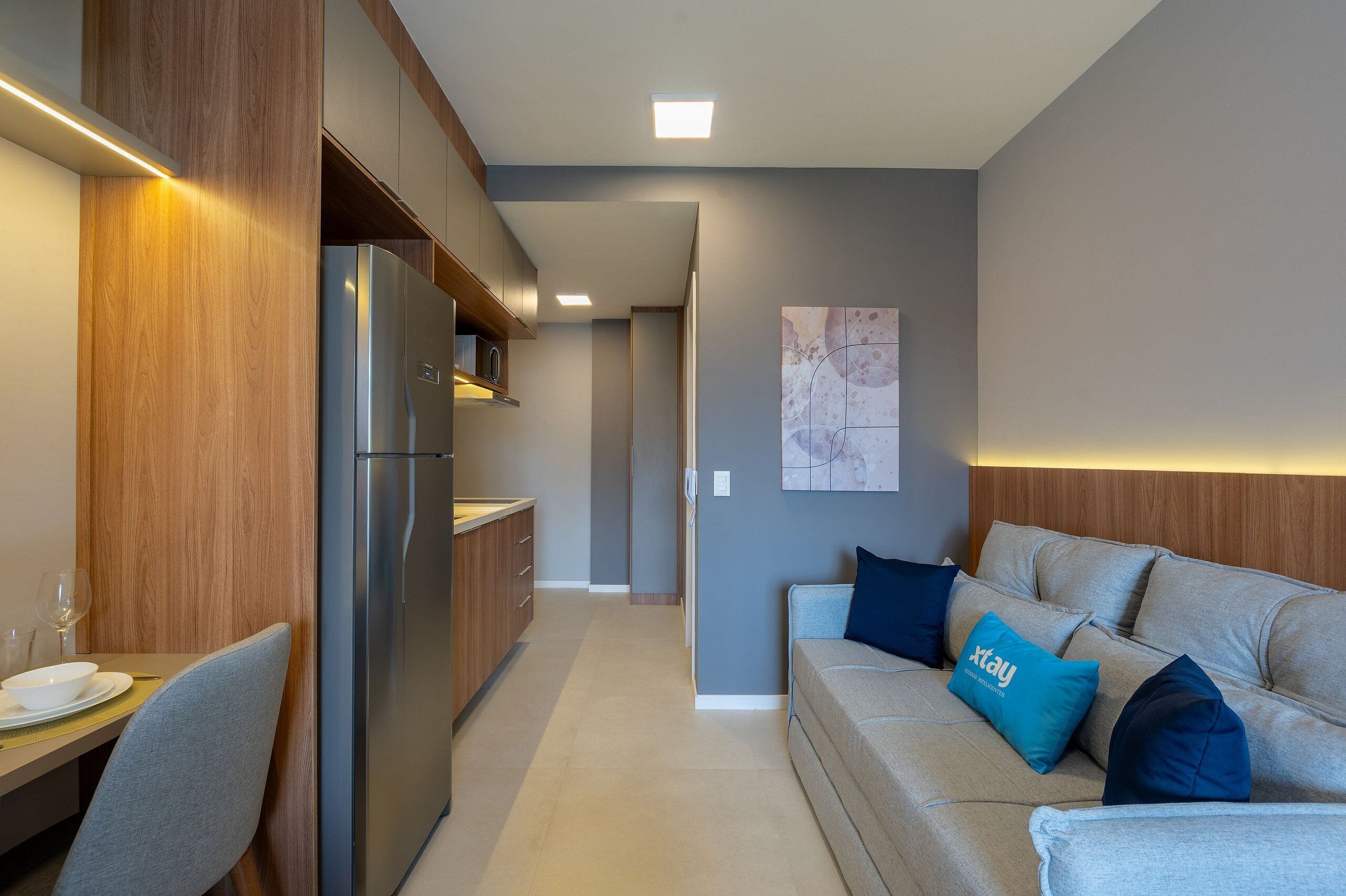 A modern kitchen with a built-in refrigerator and wooden cabinets. There is a dining area with a table and chairs, and a living area with a sofa and cushions.