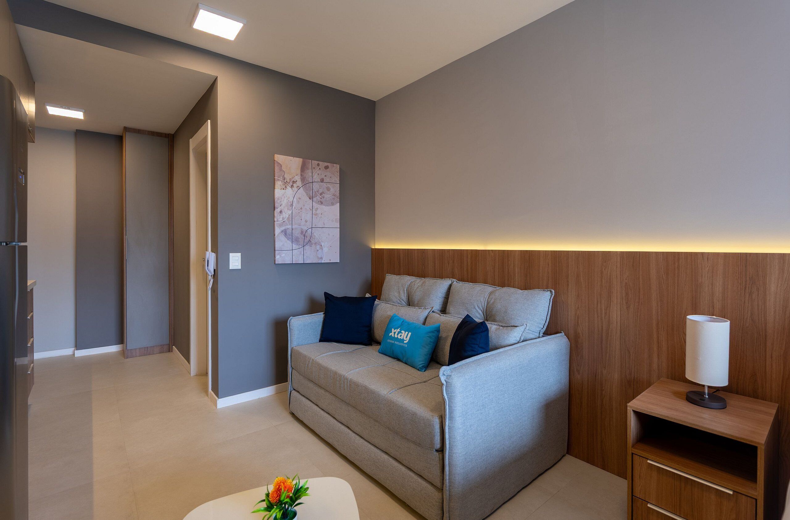 A modern living room with a sofa, a small table with a lamp, and a wall-mounted artwork.