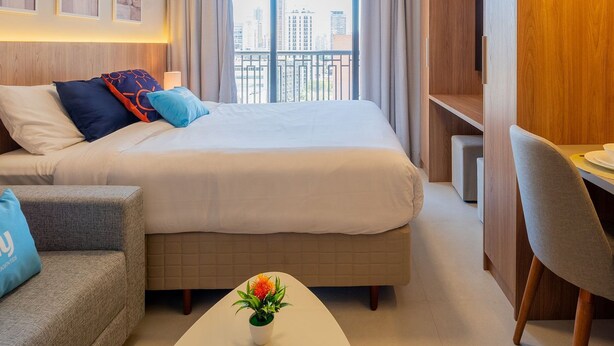 A modern hotel room with a large bed, a sofa, a small table with a plant, and a desk area. - Upper West Perdizes by Xtay (São Paulo)