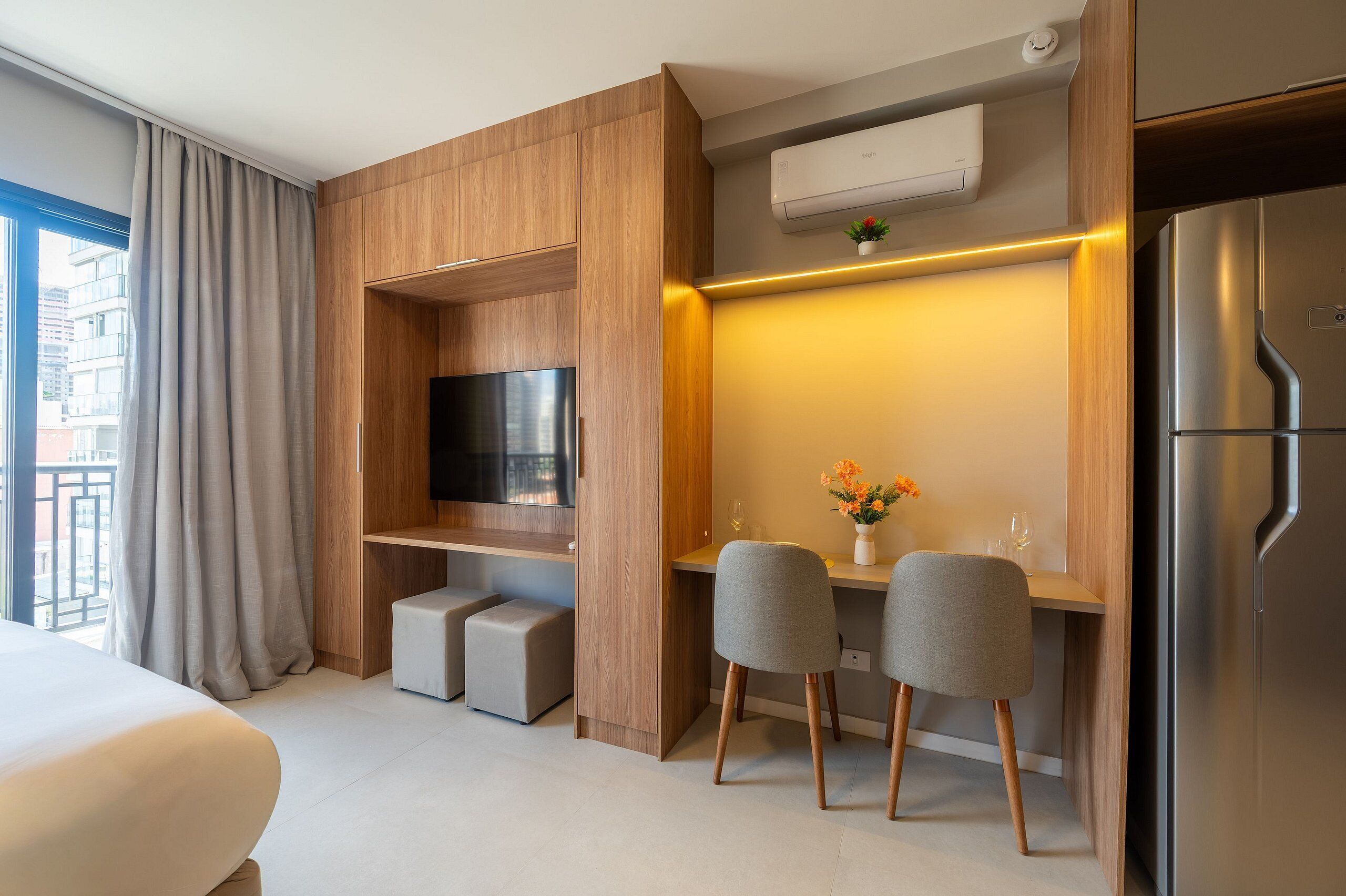 A modern hotel room with a flat-screen TV, a dining area with two chairs, a small table, and a refrigerator.