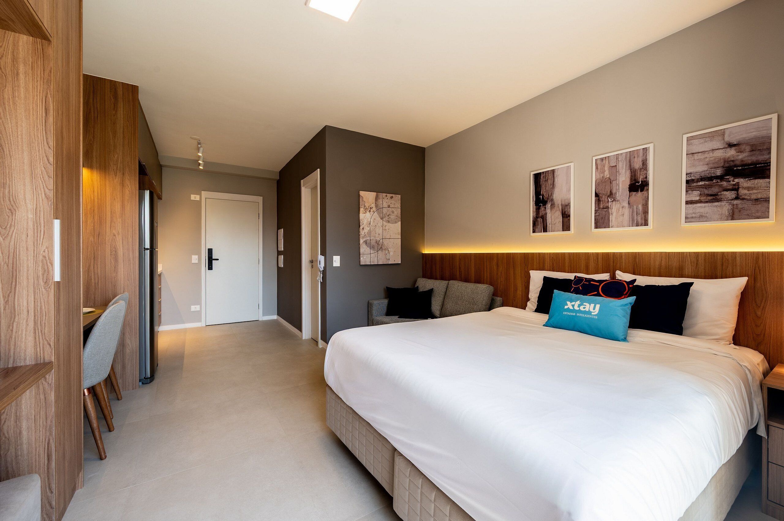 A modern hotel room with a large bed, a desk, and a chair. There are three framed pictures on the wall and a blue pillow with the xstay logo.