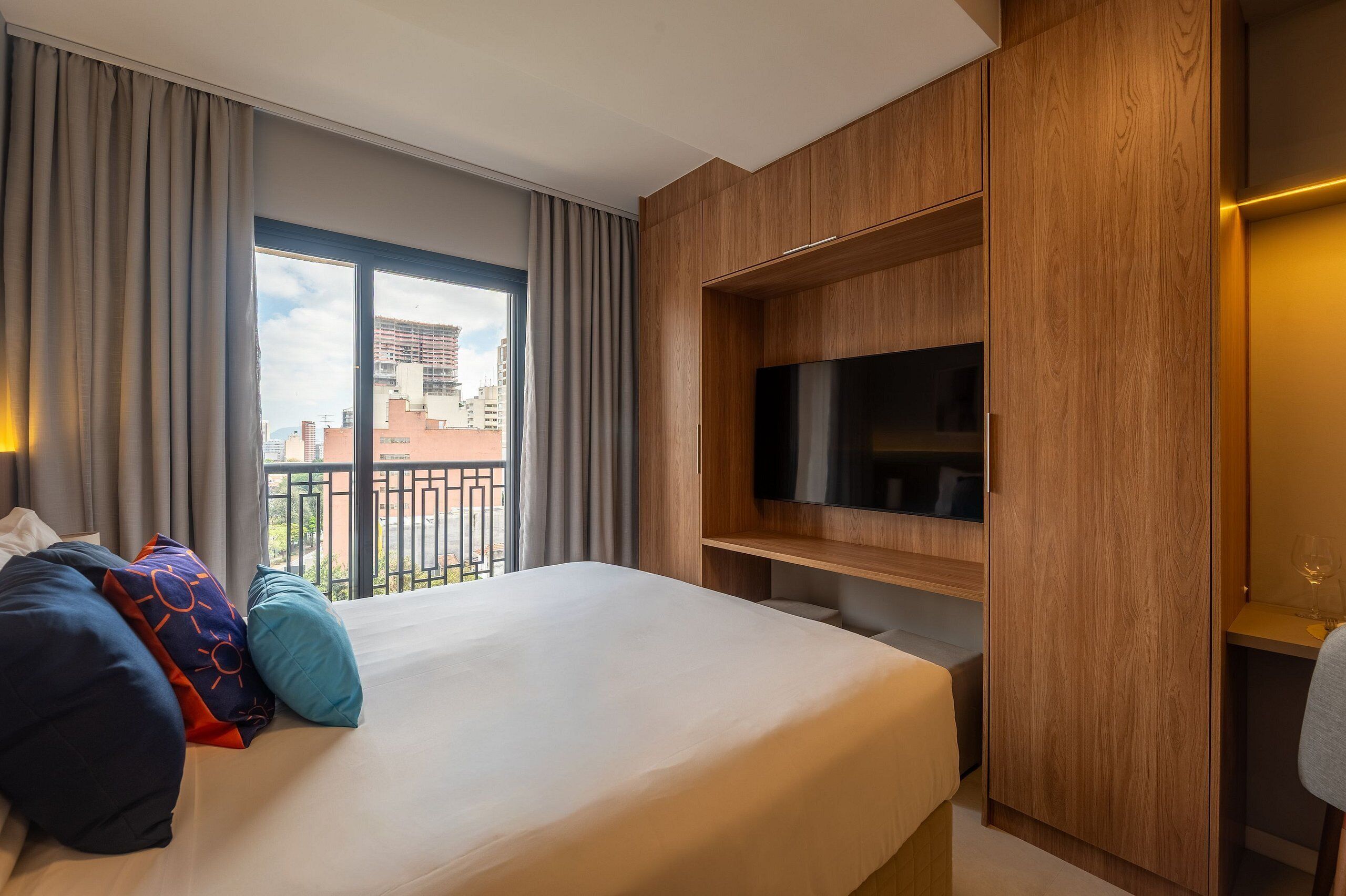 Een hotelkamer met een groot bed, een televisie bevestigd aan een houten wand en een balkon met uitzicht over de stad.