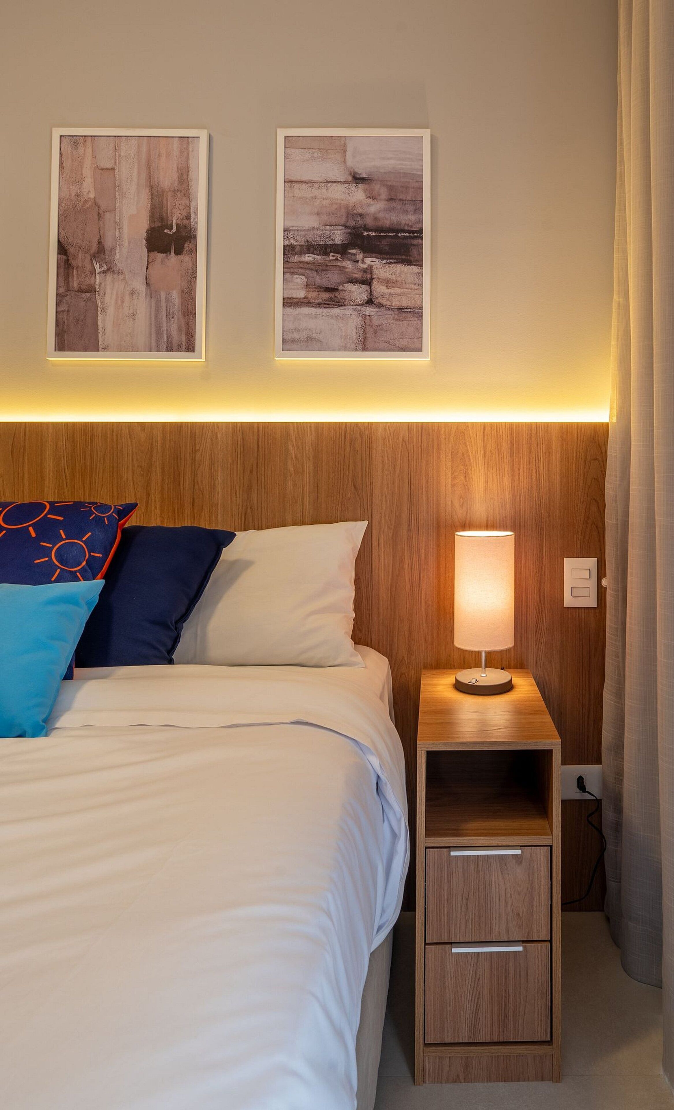 Een netjes opgemaakt bed met witte en blauwe kussens, een nachtkastje met een lamp, en twee abstracte schilderijen aan de muur.