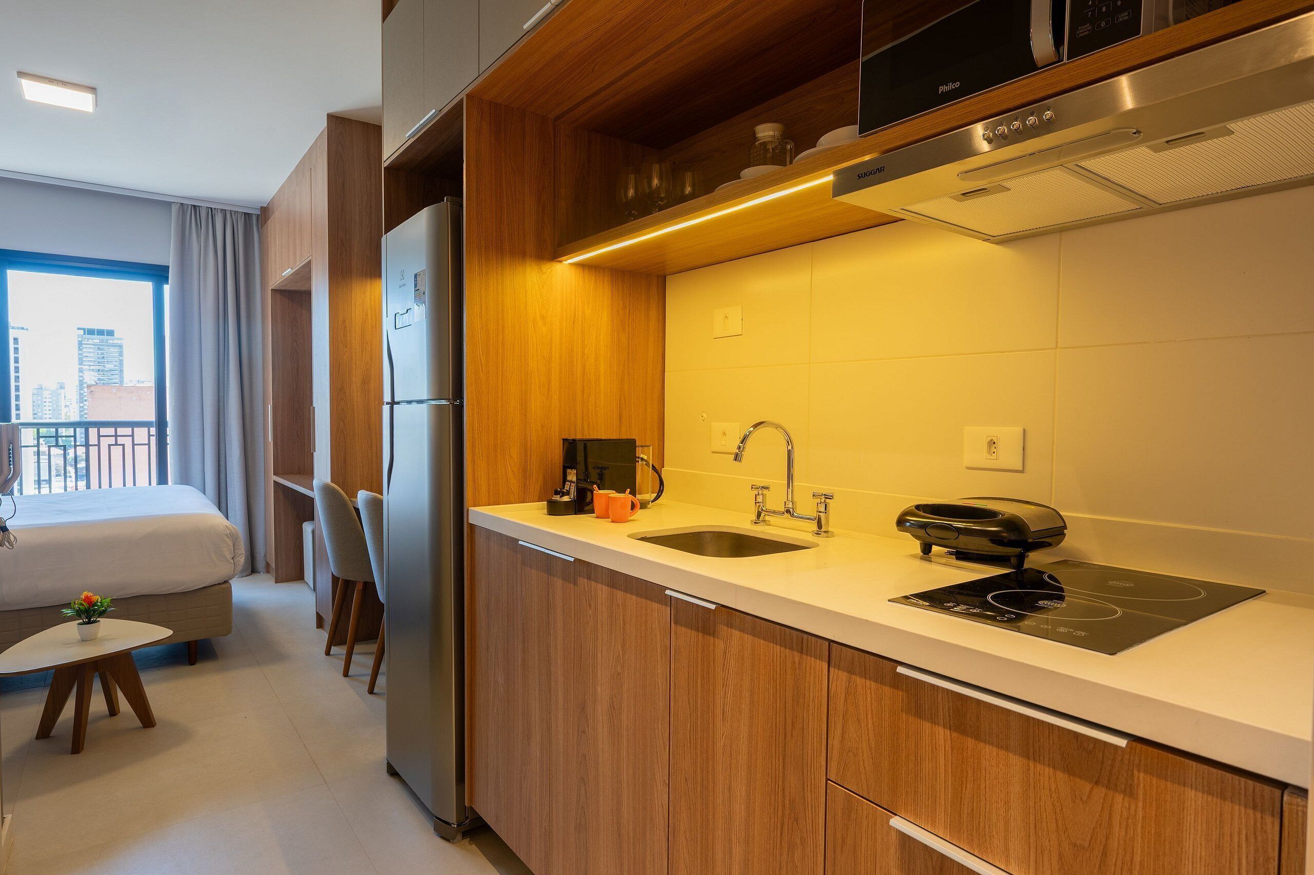 Quarto de hotel moderno com cozinha compacta, cama, uma mesinha com uma planta e vista da cidade por uma grande janela.