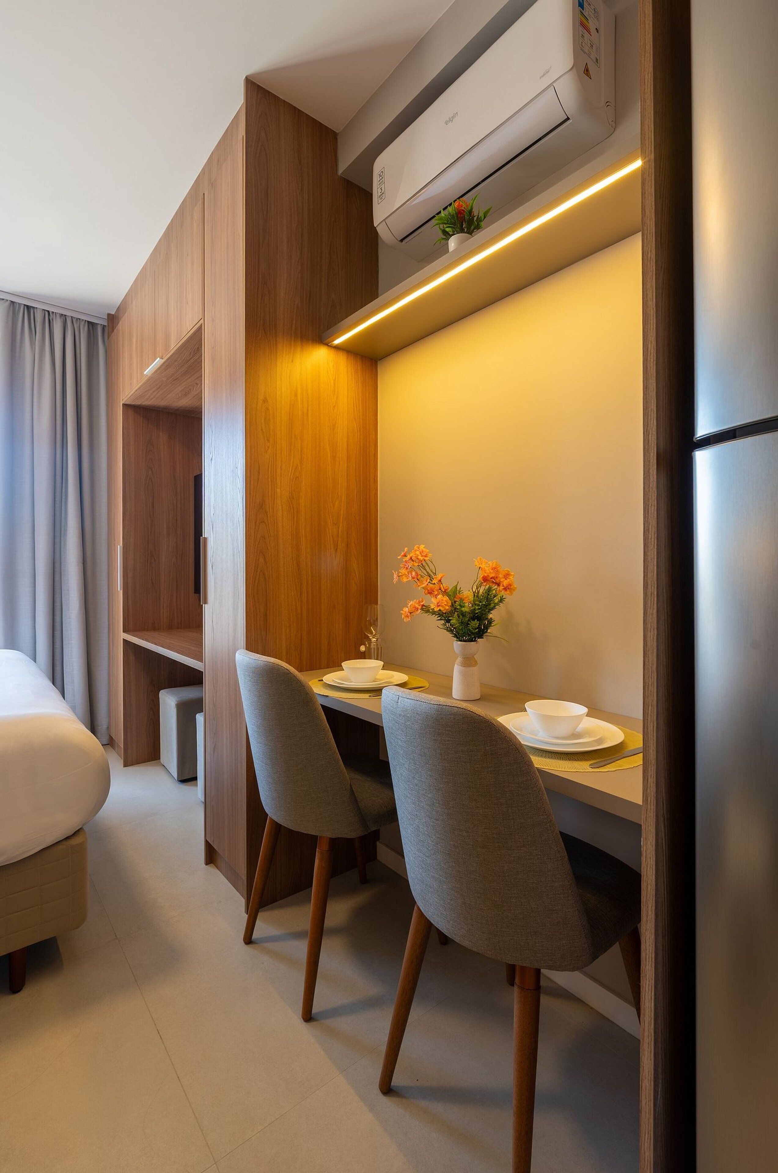 A compact hotel room with a bed, a small dining table with two chairs, a white vase with flowers, and an air conditioning unit.