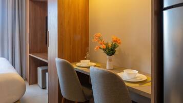 A compact hotel room with a bed, a small dining table with two chairs, a white vase with flowers, and an air conditioning unit.