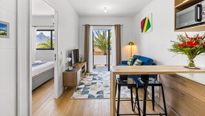 Ein kompakter, moderner Raum mit einem Bett, einem kleinen Schreibtisch, einem Stuhl, einem Fernseher und Blick auf Palmen durch ein Fenster.