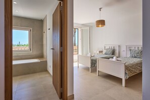 Ein Schlafzimmer mit großem Fenster, einer integrierten Sitzbank, zwei Betten mit gemusterten Betttextilien und einer hängenden Leuchte.