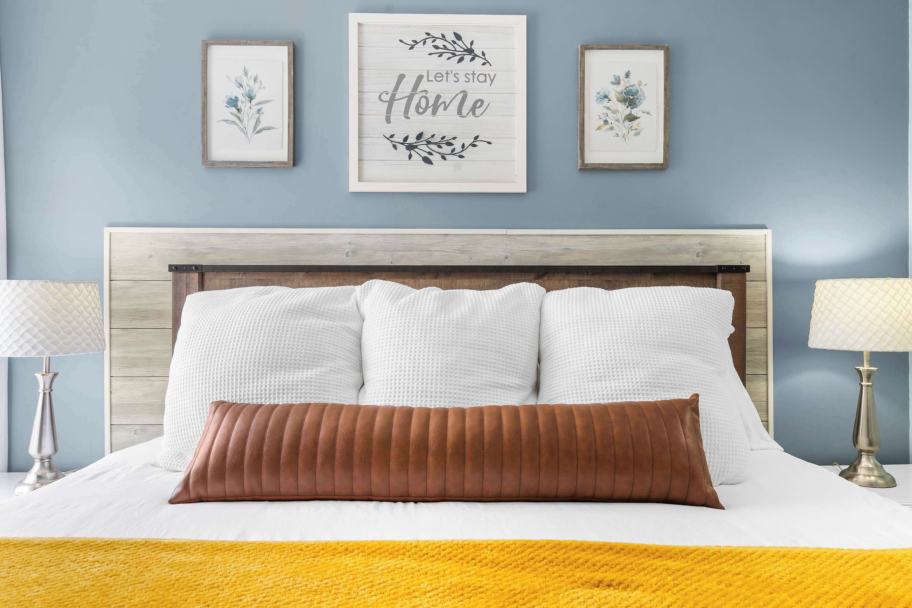 A bed with white linens and a brown pillow, flanked by two bedside lamps. Above the bed, there are three framed artworks with floral designs and a central sign that reads "Let's stay Home."
