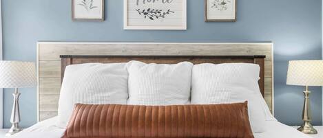 A bed with white linens and a brown pillow, flanked by two bedside lamps. Above the bed, there are three framed artworks with floral designs and a central sign that reads "Let's stay Home."