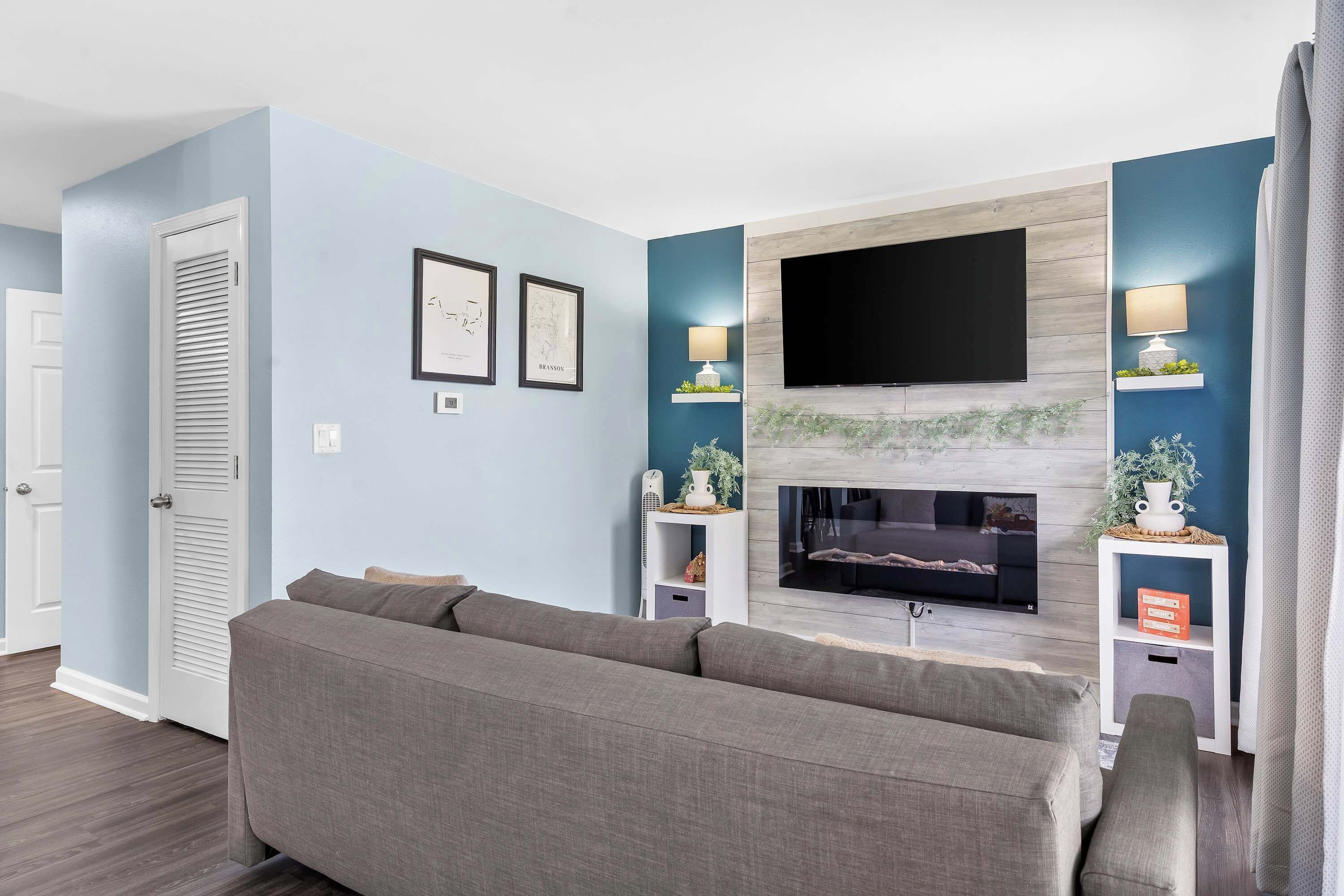 Ein Wohnzimmer mit grauem Sofa, einem Flachbildfernseher über einem Kamin und einem kleinen Beistelltisch mit Lampe.