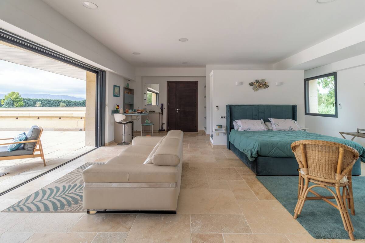 A modern bedroom with a large bed, a sofa, and a wicker chair. There is a balcony with a view of the mountains.