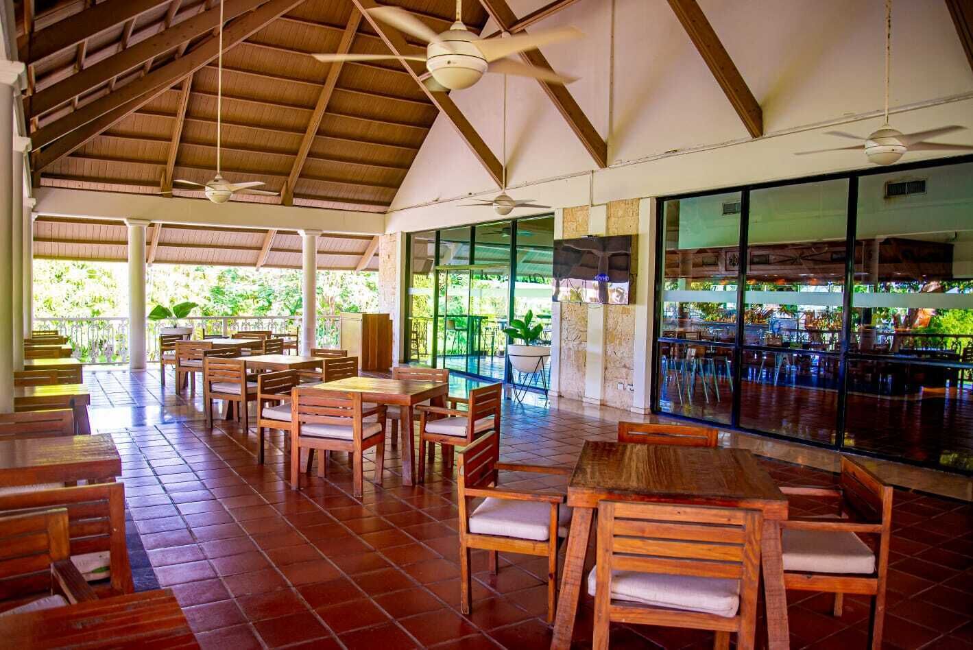 Un espacio interior amplio con mesas y sillas de madera, techo alto con vigas de madera y ventanas grandes que ofrecen vistas a la vegetación exterior.