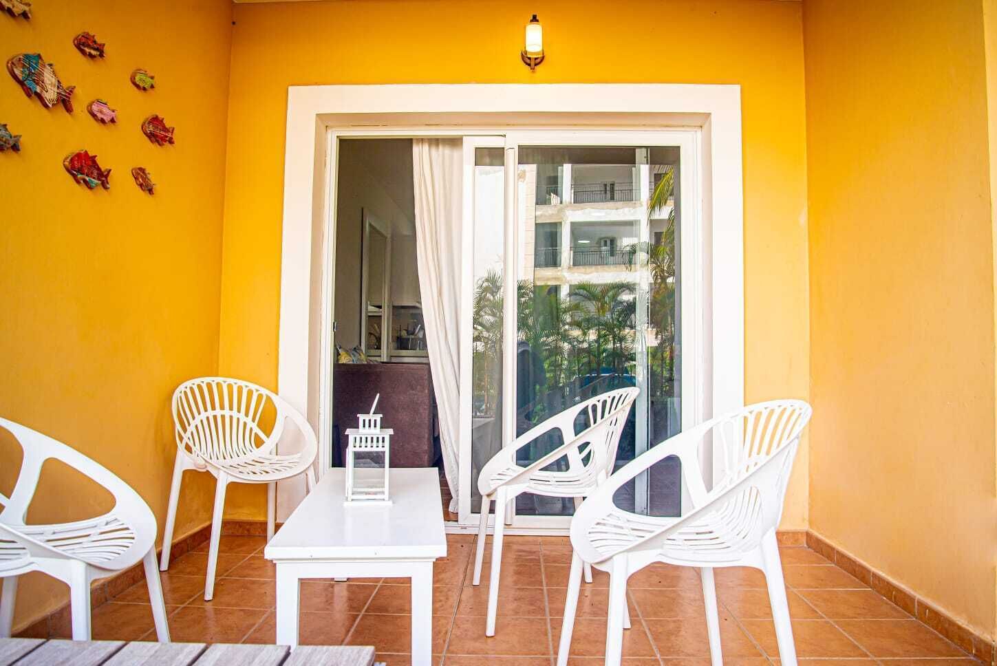 A balcony with white plastic chairs and a table, yellow walls, and a view of a building.