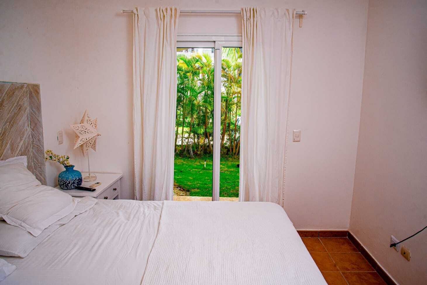 A bedroom with a bed, a nightstand, a lamp, and a window with curtains.