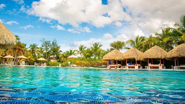 Une piscine d’eau cristalline, bordée de parasols en chaume et de chaises longues, le tout entouré de palmiers et d’une végétation tropicale luxuriante.