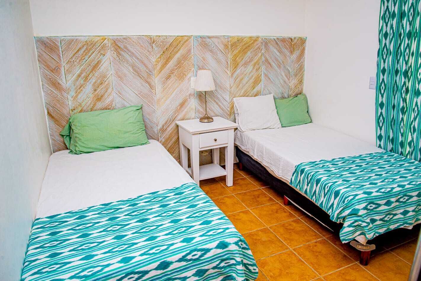 A room with two beds, each with a green pillow and a patterned bedspread. There is a small white nightstand with a lamp between the beds. The walls have a chevron pattern, and the floor is tiled.