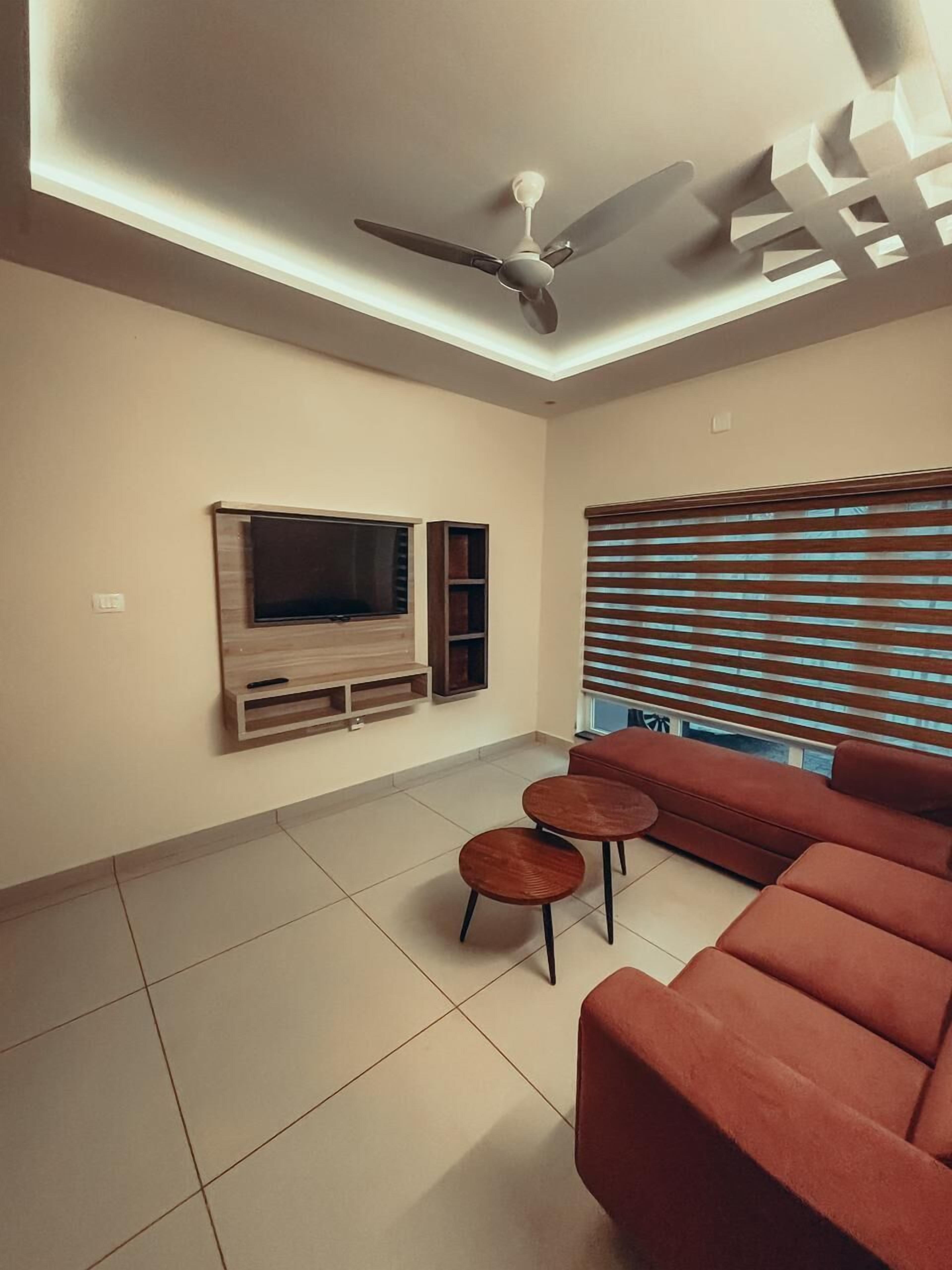 Una sala de estar con una televisión de pantalla plana montada en la pared, un ventilador de techo y un ventanal con persianas.