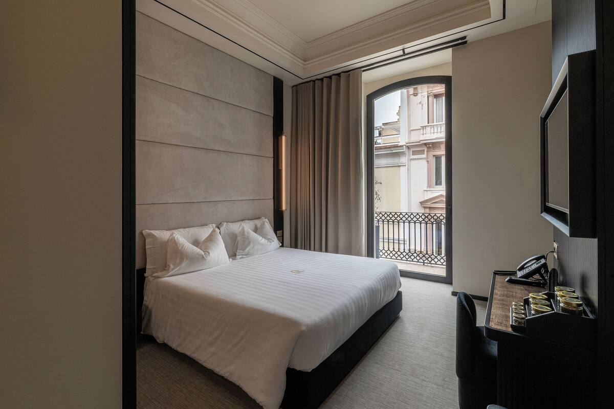 A hotel room with a large bed, a desk with a phone, and a view of the outside balcony.