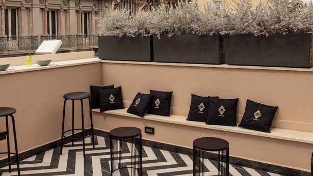 Eine Dachterrasse mit einem gemusterten Boden, schwarzen Hockstühlen und einer Sitzecke mit Kissen.