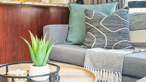 Ein Wohnzimmer mit einem grauen Sofa, einem grünen Kissen und einem gemusterten Kissen. Auf einem Tablett steht eine Pflanze in einem weißen Topf, und ein Holznachttisch mit einem Tablett.