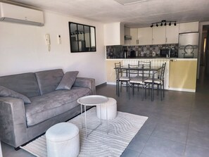 Un salon moderne avec un canapé gris, une table basse ronde et un pouf blanc. On aperçoit à l’arrière-plan un coin repas avec une table et des chaises en verre.