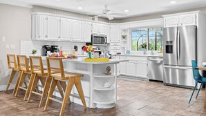 A modern kitchen with white cabinetry, a central island with bar stools, stainless steel appliances, and a view to the outdoors. - New listing discount! 5BR luxe oasis: Pickleball+bowling+golf+game room & more! (Bradenton)