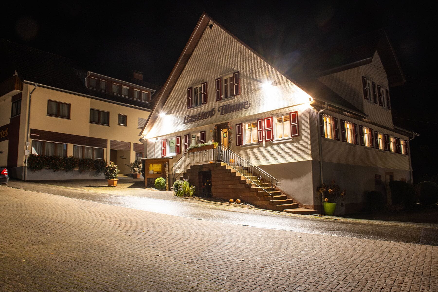Ein zweistöckiges Gebäude mit einer Treppe zum Eingang, einem Schild mit der Aufschrift 'Gasthof Blaue' und einer gepflasterten Straße davor.