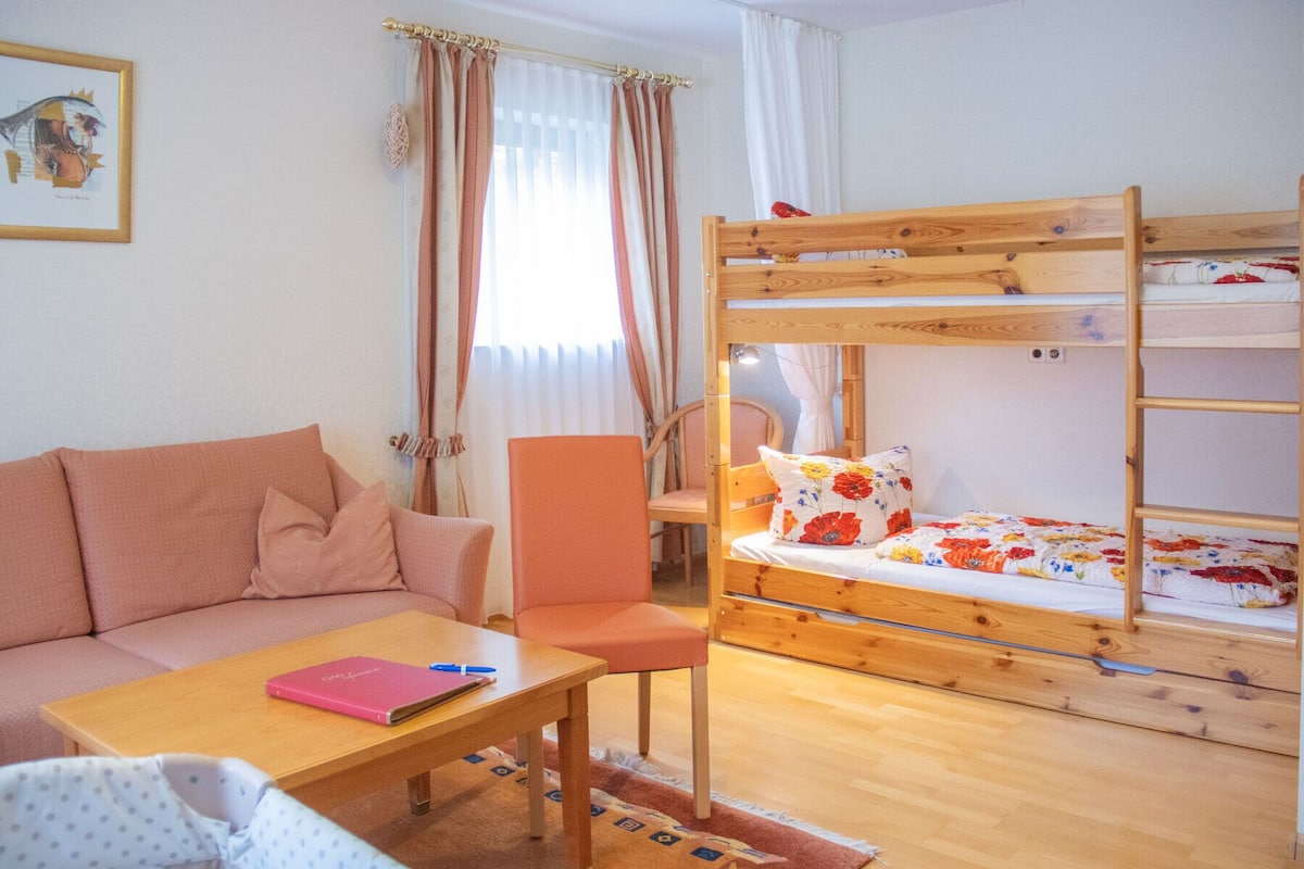 Una habitació amb un llit superposat, un sofà, una cadira, un taulell amb un llibret i una finestra amb cortines.