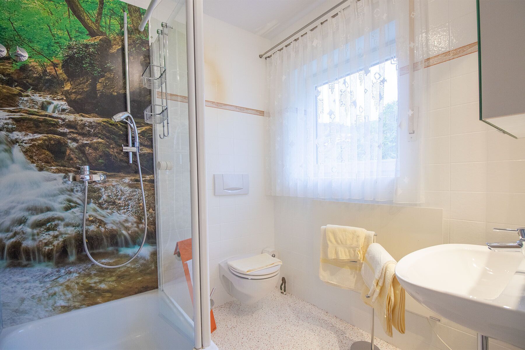 Un baño con cabina de ducha de vidrio, inodoro, lavamanos y ventana con cortinas translúcidas.