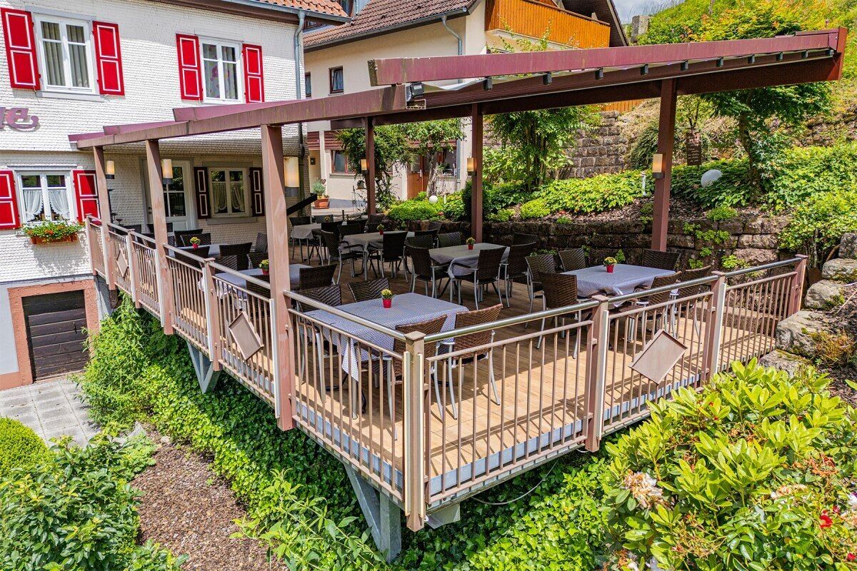 An outdoor patio with tables and chairs, surrounded by greenery and a stone wall.