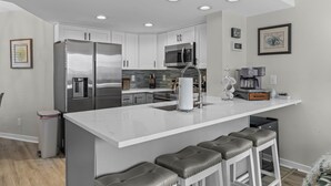 A modern kitchen with white cabinets, stainless steel appliances, and a central island with bar stools. - Port O' Call D102 - Walkout to Beach/Ocean! (Isle of Palms)