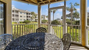 A covered outdoor area with a table and chairs, a wooden ceiling, and a view of a residential building and palm trees. - Port O' Call D102 - Walkout to Beach/Ocean! (Isle of Palms)