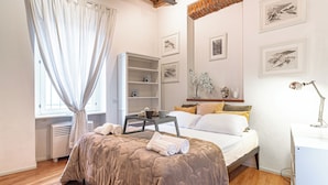 A bedroom with a bed, a bedside table, a desk, and a chair. There are framed pictures on the wall and a window with curtains. - Suite Manzoni by Houset (Monza)