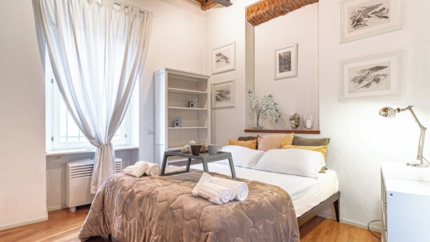 A bedroom with a bed, a bedside table, a desk, and a chair. There are framed pictures on the wall and a window with curtains. - Suite Manzoni by Houset (Monza)