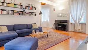 A living room with a blue sofa, a coffee table, a bookshelf, and a piano. - Suite Manzoni by Houset (Monza)