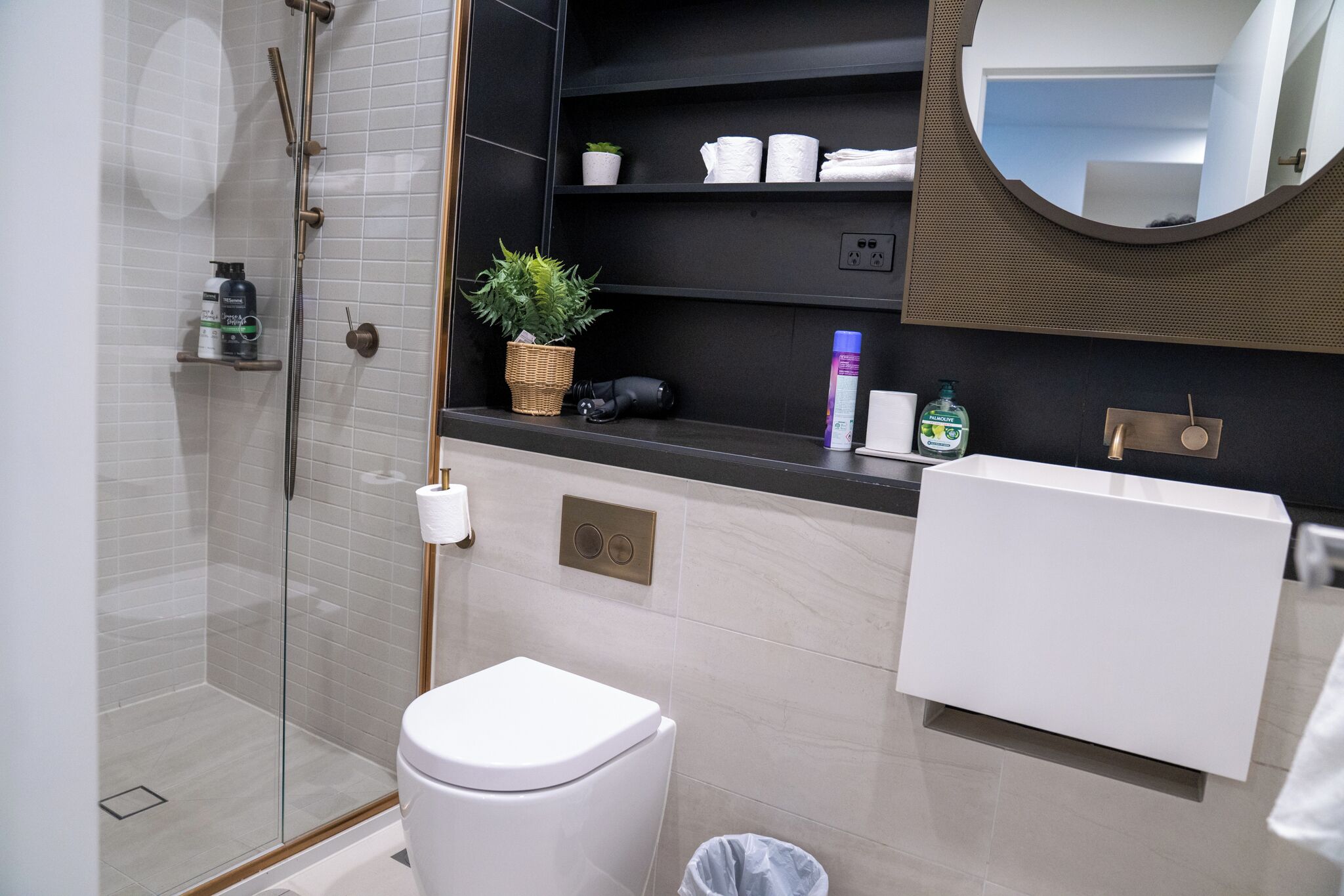 A modern bathroom with a glass shower enclosure, a white toilet, a sink with a mirror above it, and shelves with toiletries.