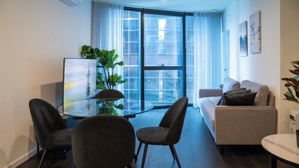 A modern living room with a glass dining table, two dark chairs, a beige sofa, and a large window with sheer curtains. - Home in the Sky 2BR,2BA with City Views,Pool & Gym (Melbourne)