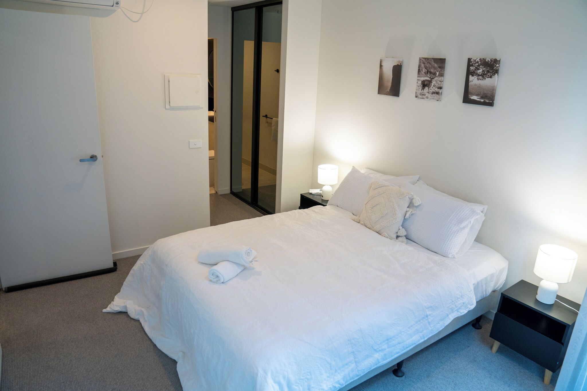 Un lit bien fait, avec du linge de lit blanc et deux serviettes enroulées, une table de chevet avec une lampe, et un mur orné de trois tableaux encadrés.