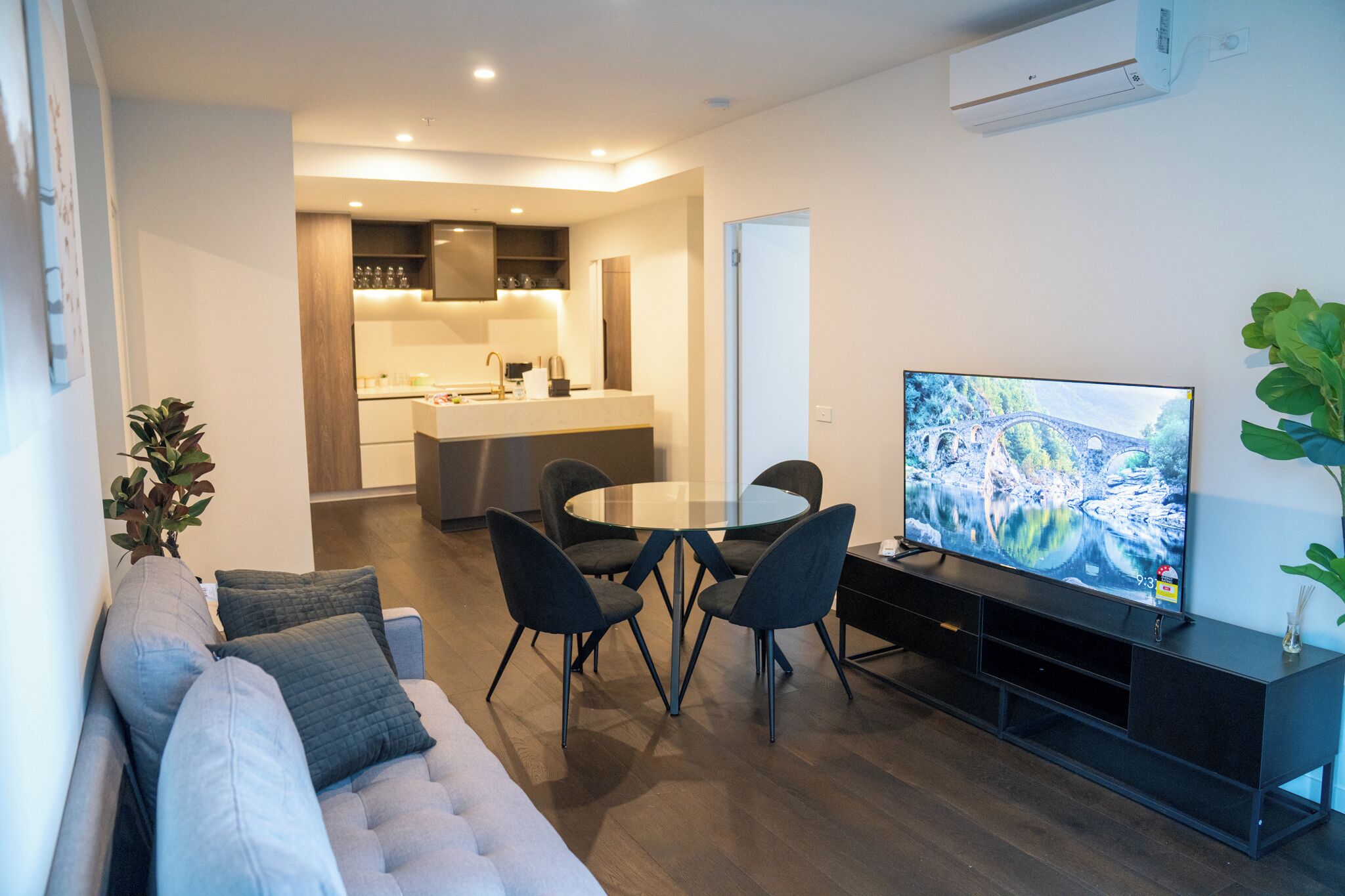 Ein modernes Wohnzimmer mit einer Couch, einem runden Glastisch und vier Stühlen. Ein Flachbildfernseher ist an einer schwarzen Unterhaltungseinheit befestigt.