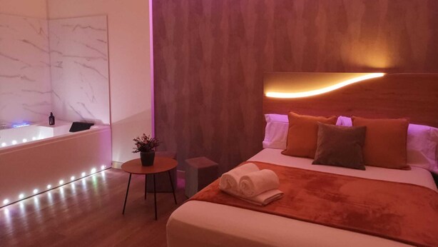 Quarto de hotel moderno com uma cama grande, uma pequena mesa com uma planta, uma luminária na parede e um banheiro com banheira e iluminação.