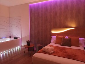 A modern hotel room with a large bed, a small table with a plant, a wall-mounted light, and a bathroom with a bathtub and lights. - Paradise Suites Madrid (Madrid)