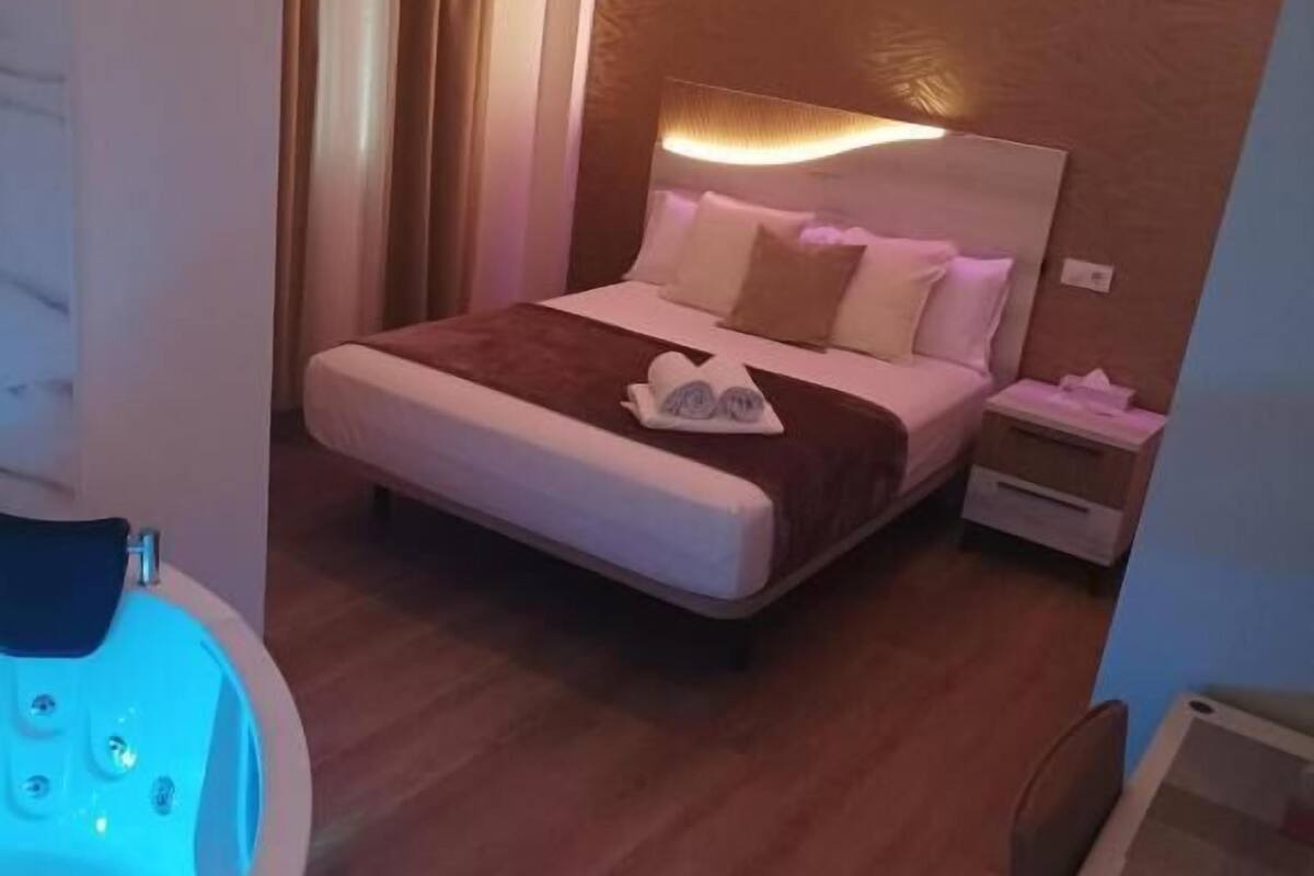 A hotel room with a large bed, a bedside table, a nightstand, a bathroom with a bathtub, and a wall-mounted heater.