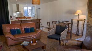 Un salon avec un canapé en cuir, une table basse en bois et un fauteuil en osier. On y trouve une table à manger avec des chaises et une lampe. Une cuisine est visible au fond.