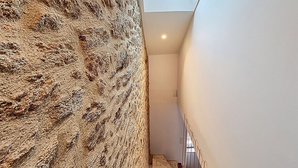 Un couloir étroit, avec un mur texturé d’un côté et un mur blanc de l’autre, qui mène à un escalier surmonté d’une lucarne.