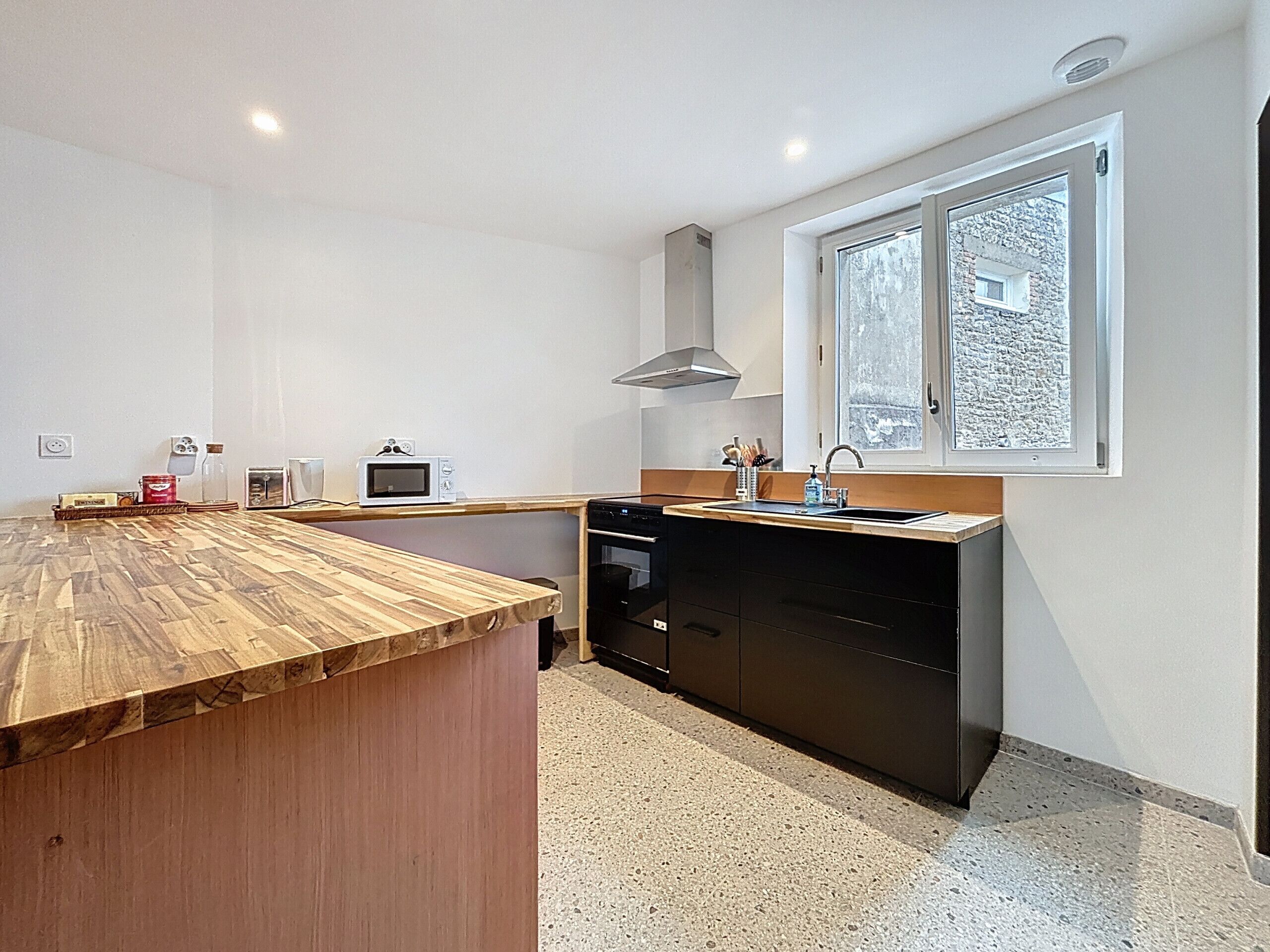 A compact kitchen with a wooden countertop, a microwave, a sink, and a window with a view of a stone building.