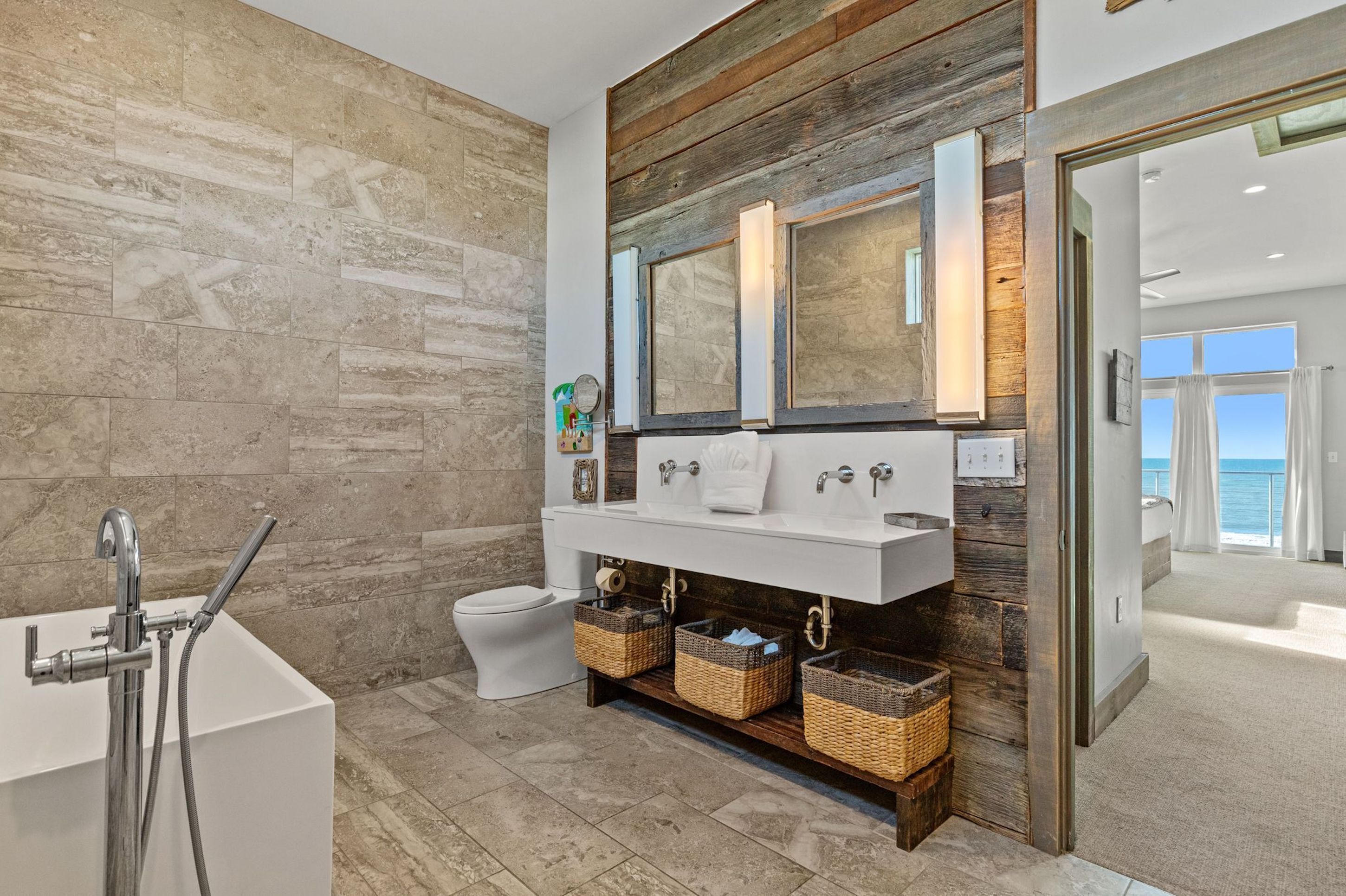 Een moderne badkamer met een stenen wand, een houten accentwand, een grote spiegel, een witte wastafel, een toilet en rieten manden onder de wastafel.
